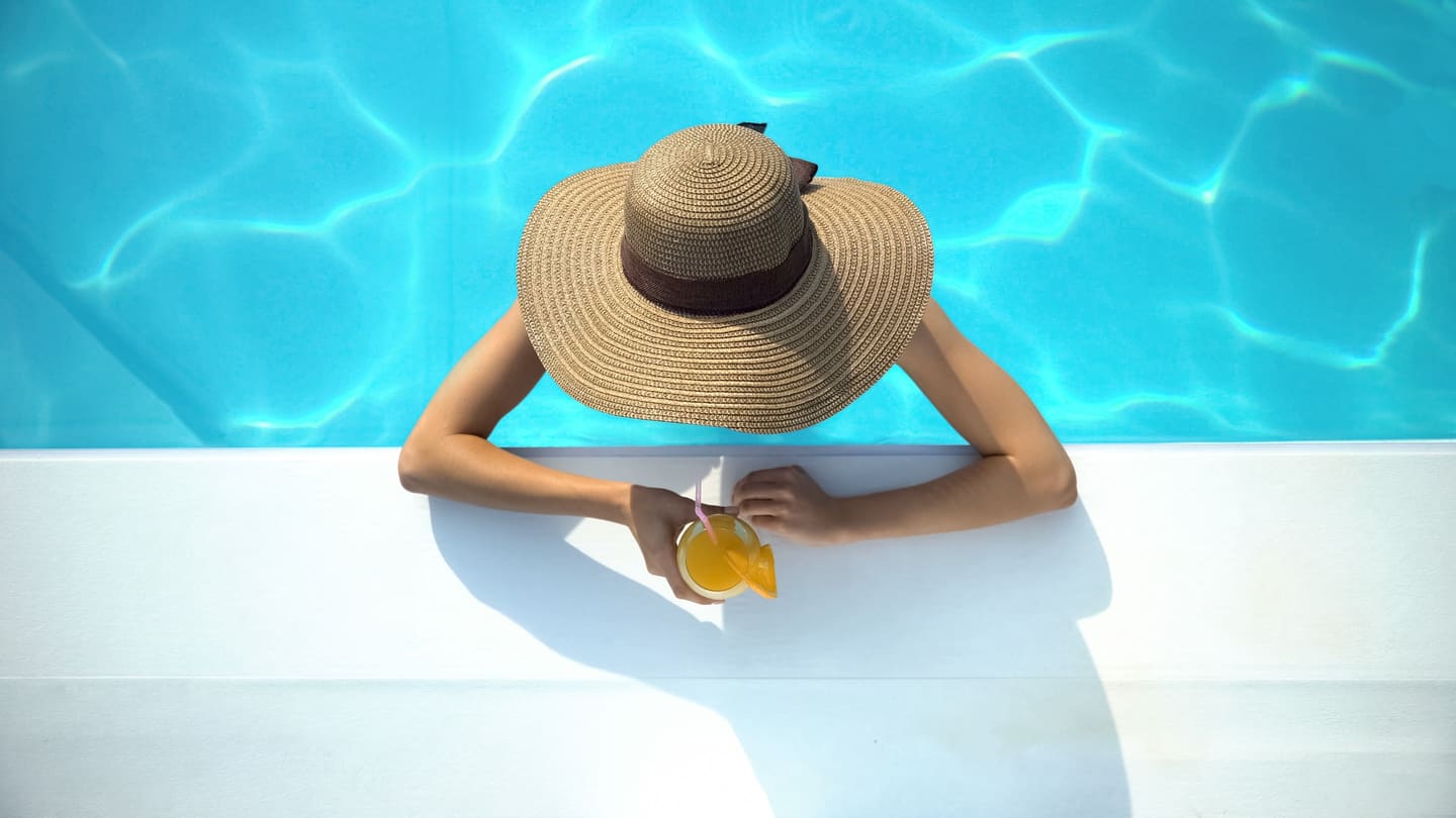 A woman in a straw hat resting at the edge of a turquoise villa pool with a drink